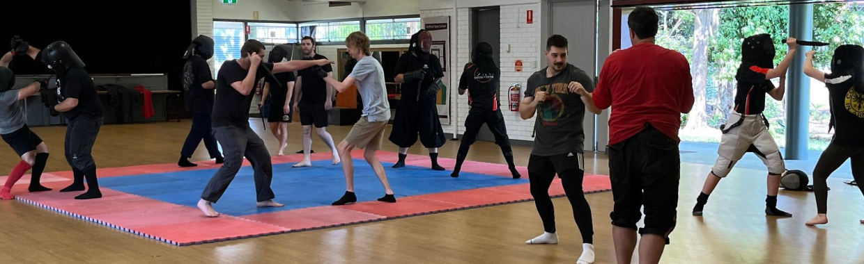 A room full of people practicing swordplay techniques in pairs.